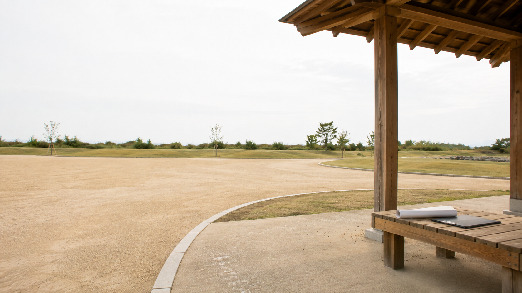 公園の四阿の下に置かれた木製ベンチと、その上のクリップボードと巻いた図面